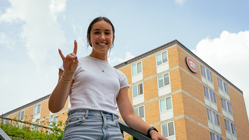 Female student leaving campus dorm room and giving the "sign of the husky"