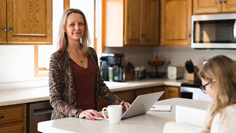 A mom works on her laptop at a kitchen island alongside her child