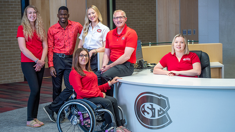 A small group of SCSU employees gathered around a reception desk, posing for a photo. One employee is seated in a wheelchair.