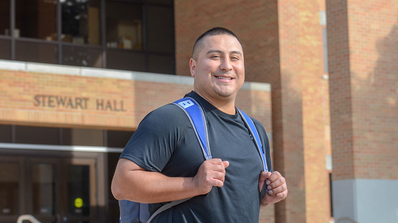 Older male student stands outside of Stewart Hall with backpack
