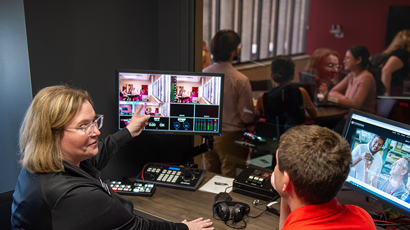 Two people observe students in the Sales Lab via live camera footage on a computer screen