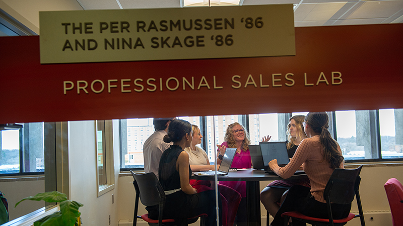 From outside of the Professional Sales Lab, a staff person speaks with several students around a high top table