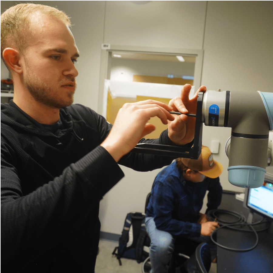 Student adjusting equipment in an indoor laboratory while another student sits nearby