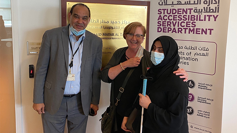 Dr. Kathy Johnson standing in between a blind female with a cane wearing a dress with a scarf and a guy in a business suit. Background has a tan wall and an advertisement promoting disability rights. 