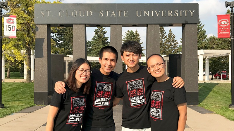 Group photo of 1 Asian woman and 3 Asian men standing in front of a dark granite pillars with "St. Cloud State University" embedded above the pillars. The background has blue sky, green grass, and pine trees. 