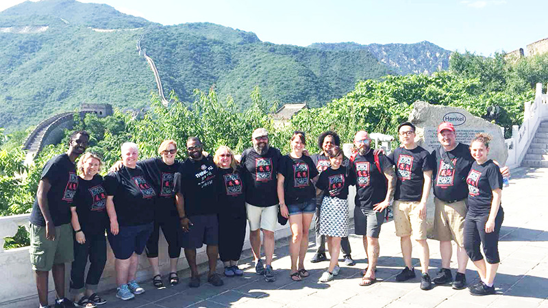 Group photo on top of the Great Wall consisting of 14 people of both genders, all wearing black Confucius t-shirts. The background has vivid green forest surrounding the Great Wall from all around.