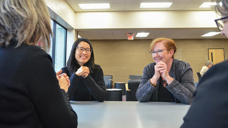 Two women smiling and chatting at a table with two other women with their back to the camera. Left Asian woman with glasses, right redhead woman with glasses. Brick walls in the background.