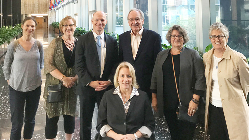 Group picture of an attendee in wheelchair, Dr. Kathyn Johnson, Dr Amy Knopf, former senator Tom Harkin and colleagues at the US State Department during the United Nations for a Disability Event with vibrant flags showcasing a variety of colors from different countries above them. 