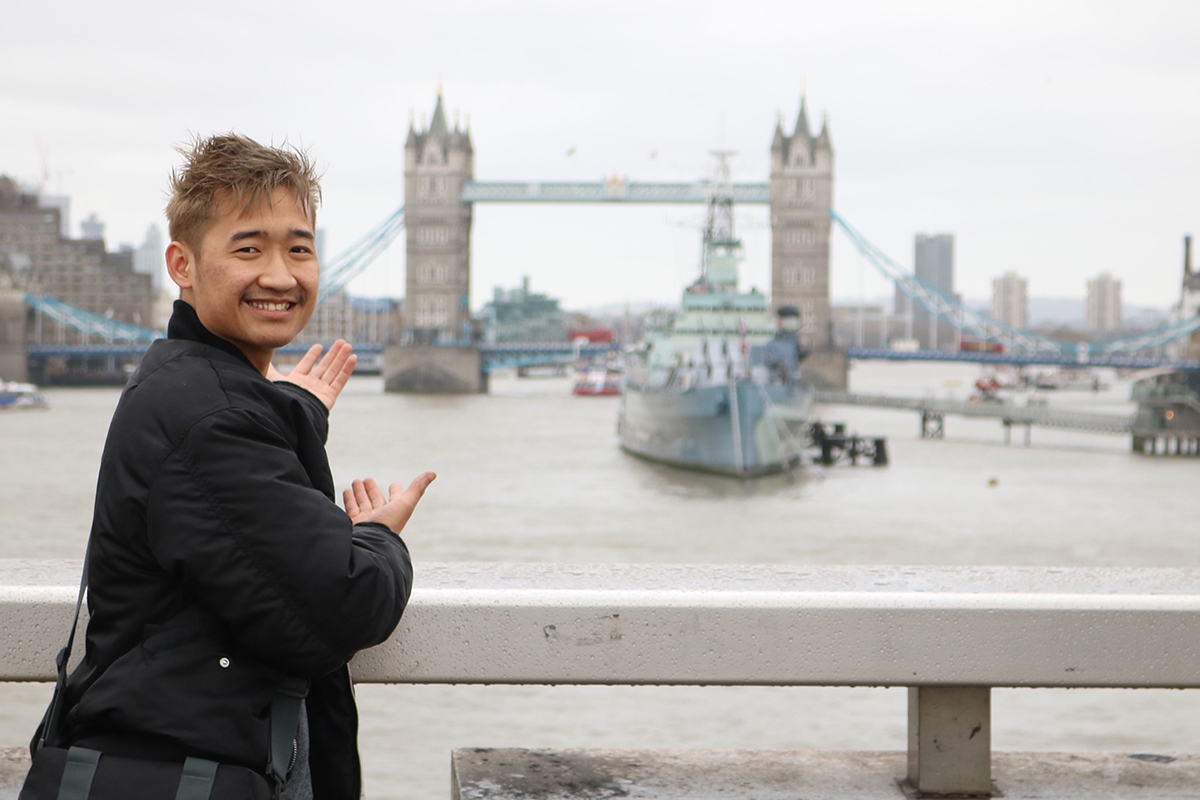 A person stands by a riverside railing, gesturing toward Tower Bridge across the River Thames, with a large ship visible on the water in the background