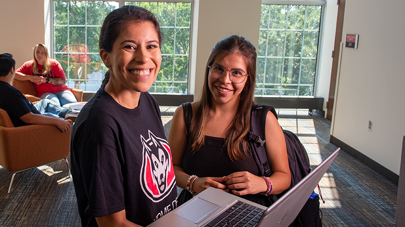 Two smiling students standing in sunny lobby area with laptop open