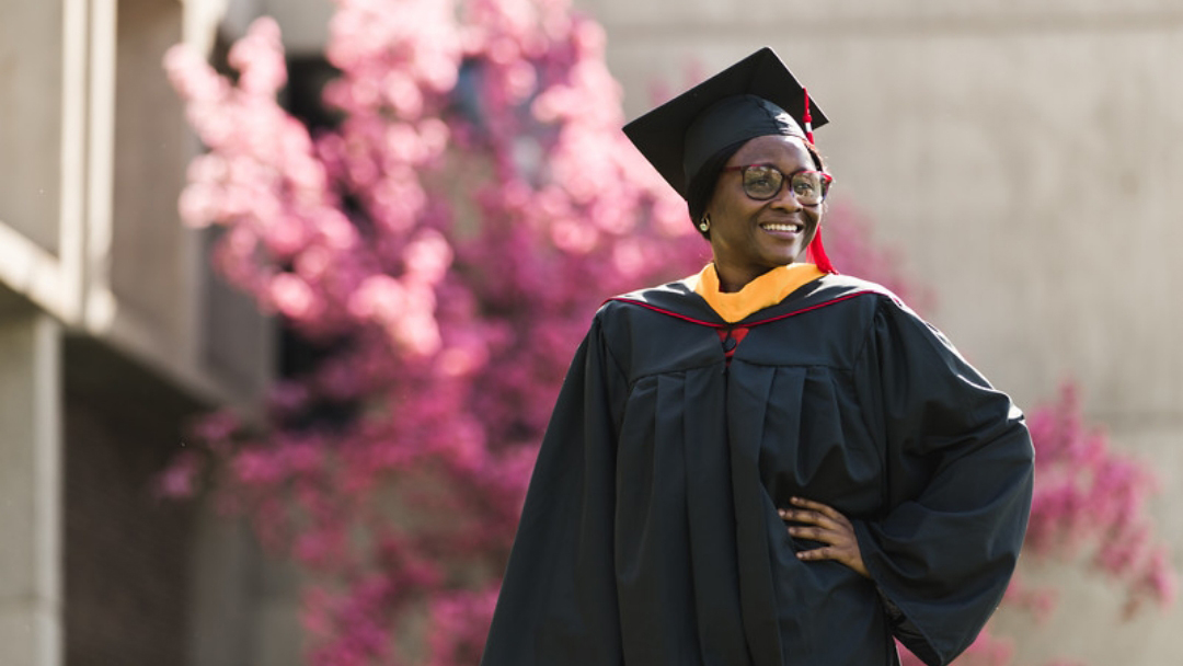 A graduate smiles with full regalia outdoors in front of blossoming tree