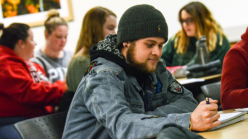 A student seated in a classroom writing with a pen on notebook paper, with other students visible in the background gathered at desks