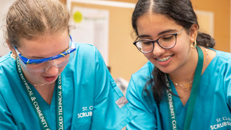 Two high school students wear blue scrubs and practice a nursing technique at Scrubs Camp
