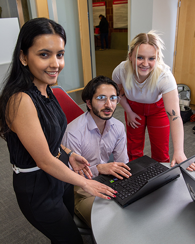 Three business students gathered around laptop, smiling at camera