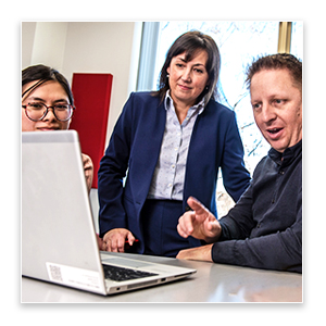 Three people gathered around a table reviewing employment information on an open laptop in an office setting.
