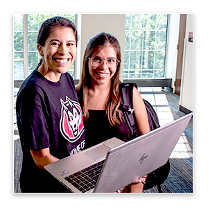 Two people standing indoors and looking at an open laptop, with large windows and natural light behind them.