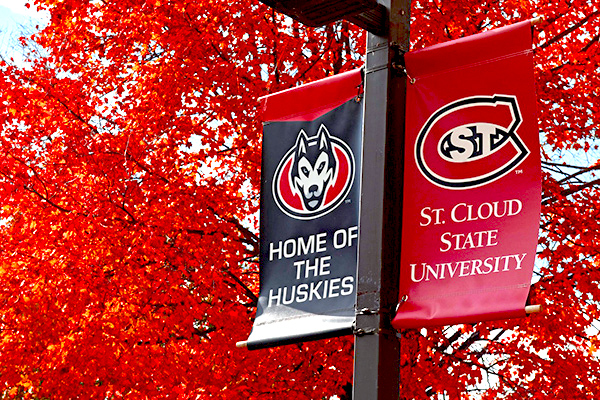 "Home of the Huskies" and "St. Cloud State University" banners hung on campus light poles in front of a tree with bright red leaves