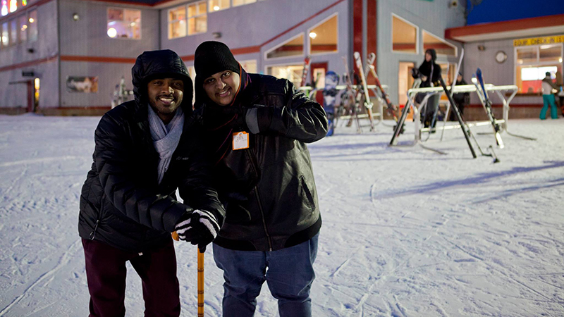 Two people bundled in winter clothing smile and pose together at a snowy ski area with warm lights from a nearby building in the background.