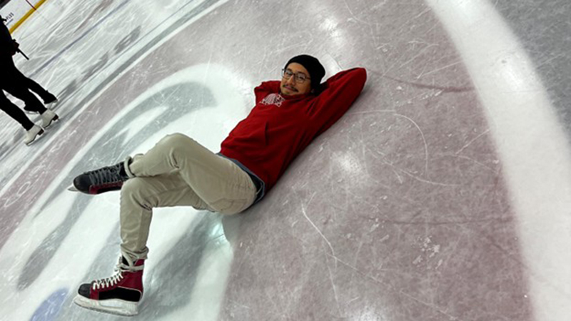 A person wearing ice skates and a red sweatshirt is lying on their back on an indoor ice rink while others skate in the background.