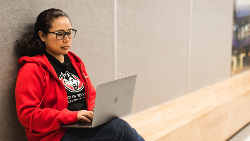 Person wearing a red zip-up sweatshirt typing on a laptop while sitting down