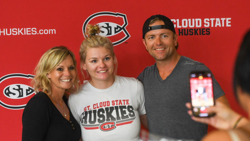 Three people having their photo taken on a cell phone in front of red backdrop with SCSU logos 
