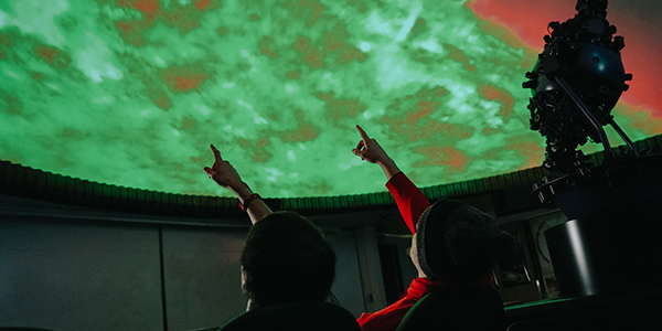 Two hand pointing up at planetarium screen showing a galaxy