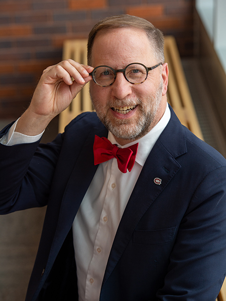 Dr. Gregory Tomso seated on a bench, adjusting his eye glasses and smiling at the camera