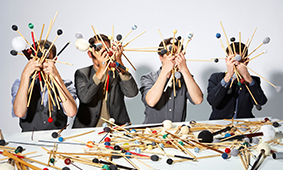 Artists hold drum sticks in front of their faces