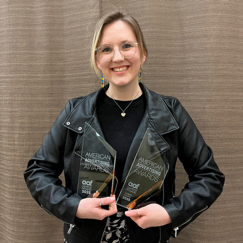 Jess Meichsner, a woman in a black jacket with glasses, holds two American Adversiting Awards while smiling