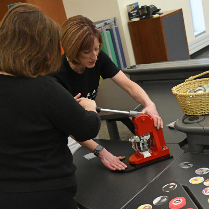 A woman demonstrates how to use a red device that makes coasters while another woman with her back to the camera watches