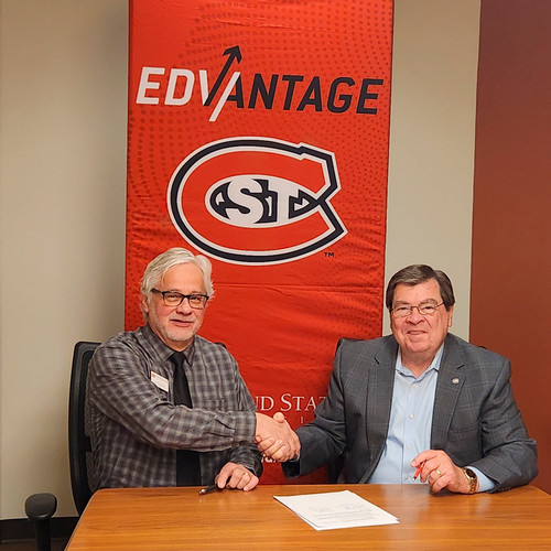  Presidents from Ridgewater and SCSU shake hands with an SCSU banner logo behind them. Both men are sitting at a table with a signed document in front of them.