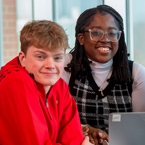 A man in a red sweatshirt and blonde hair and a woman with black hair, glasses, and a sweater smile for a photo 
