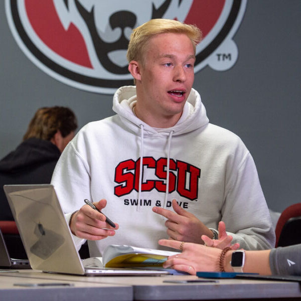 A man in a white SCSU Swim & Dive sweatshirt talks to a woman in a grey quarter zip at a table with laptops visible