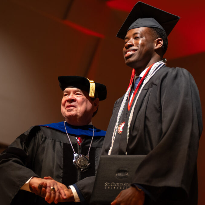 A gradaute in cap and gown smiles at the camera while shaking hands with a man in regalia