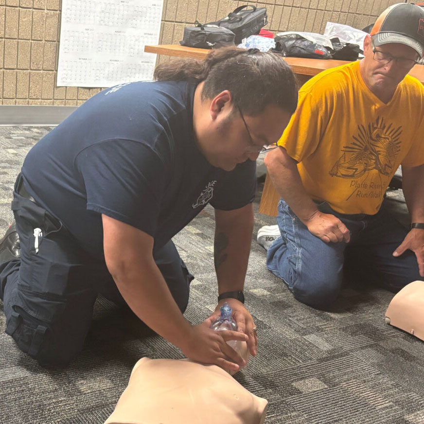 A man in a blue shirt and glasses works on a CPR mannequin while a man in a yellow shirt, glasses and hat watches in the background