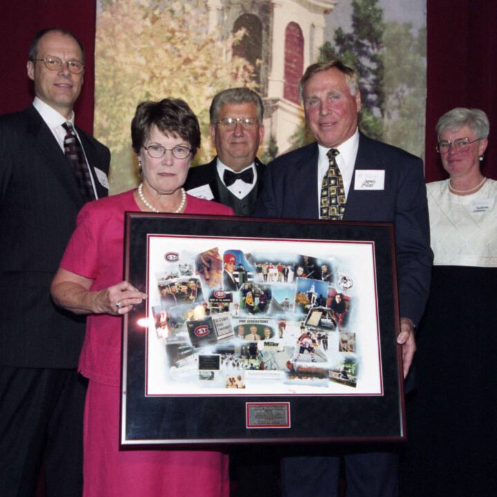 A woman in glasses and a pink dress stands next to a man in a blue suit and tie as they hold a large frame with a collection of photos. Three individuals and the Riverview cupola are visible in the background
