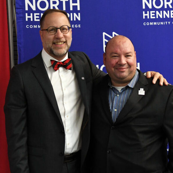 Two men in suits smile for a photo in front of logos for St. Cloud State and North Hennepin Community College