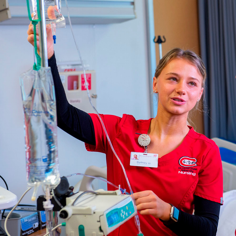 A woman in red SCSU nursing scrubs stand next to a machine in a nursing simulation lab