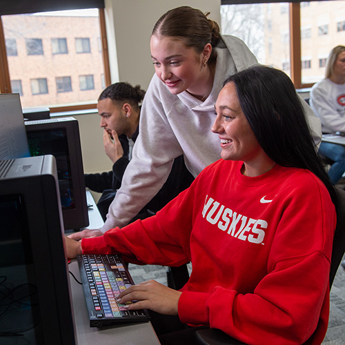 Two women look at a computer screen while students work in the background