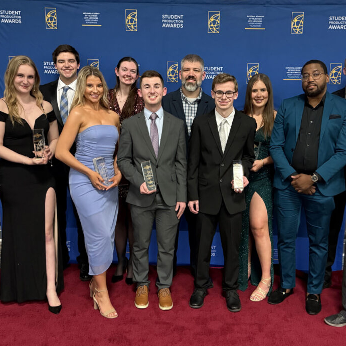 A group of nine people smiles at the camera while some hold crystal pillar awards