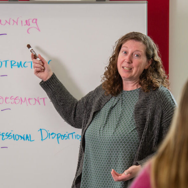 A woman in a grey sweater points to a whiteboard with a class of students out of focus
